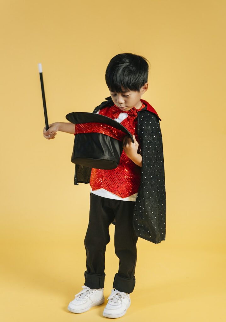 Full body of adorable magician Asian boy wearing cape standing on yellow background with wand and looking into black hat in studio