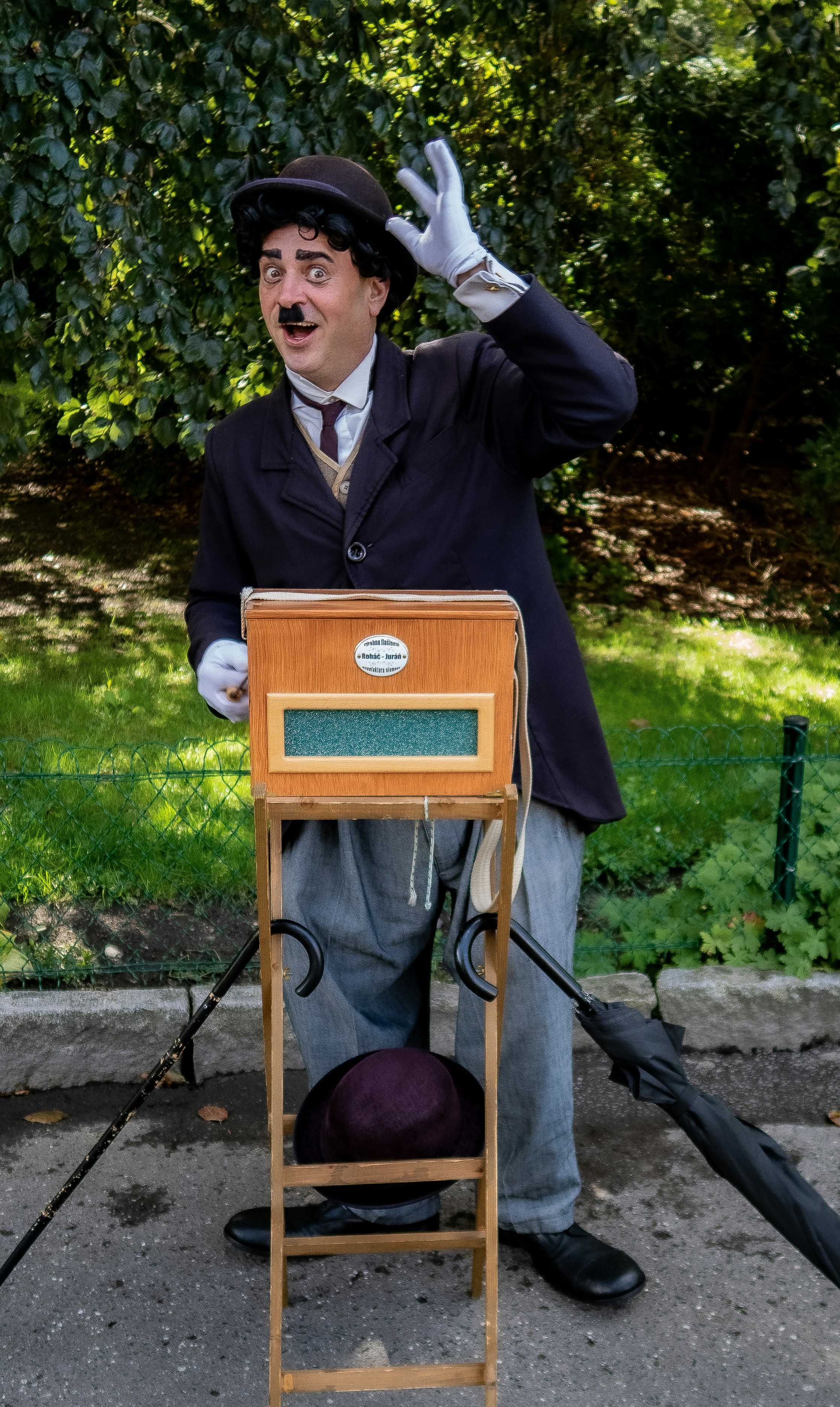 Chaplin is alive by Magic Trick man wearing Charlie Chaplin costume standing while waving his left hand near green field surrounded with tall and green trees