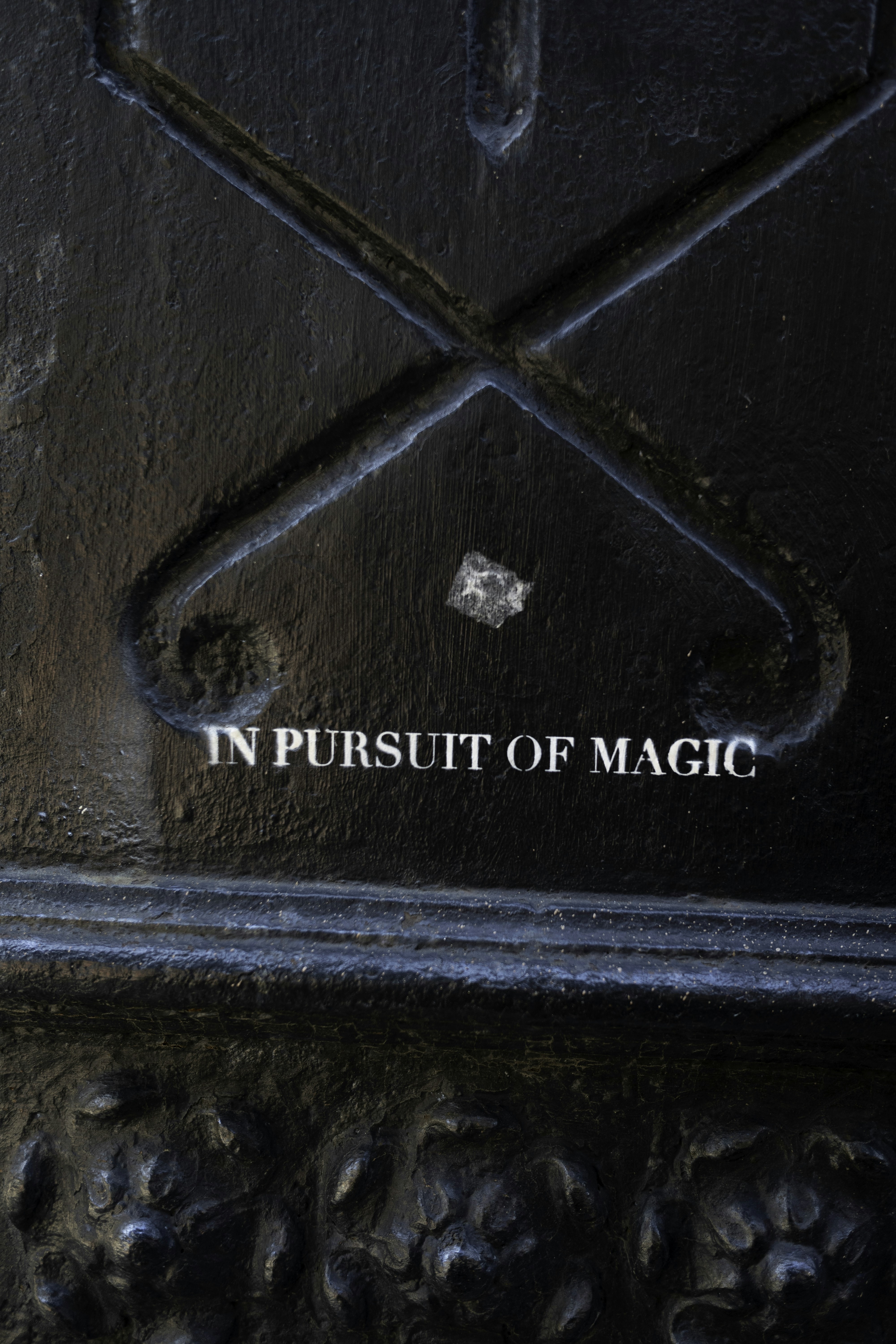 The phrase “IN PURSUIT OF MAGIC” appears in white serif lettering on a dark cast-iron surface, framed by decorative metalwork and a moody black-on-black urban backdrop. by Magic Trick Text reads
